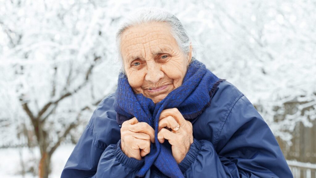 Elderly woman bundled in a blue scarf and winter coat, smiling outdoors on a snowy day — representing senior winter safety and warmth.